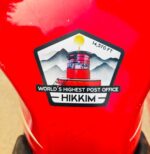 Hikkim 14,570 FT post office sticker stuck onto a red motorcycle fuel tank.
