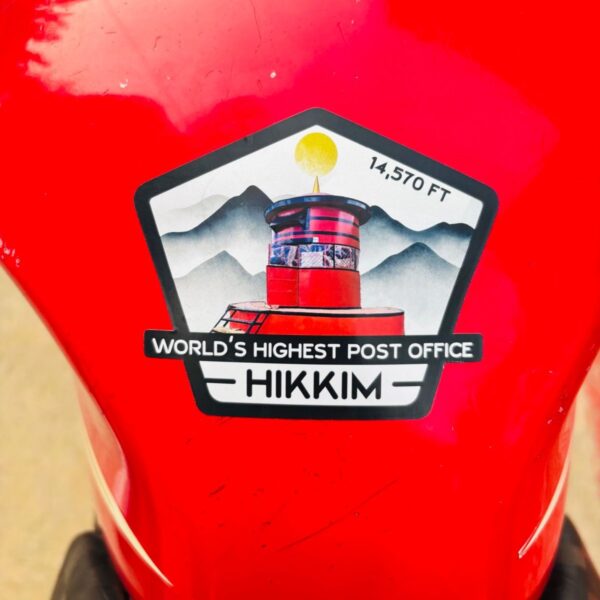 Hikkim 14,570 FT post office sticker stuck onto a red motorcycle fuel tank.