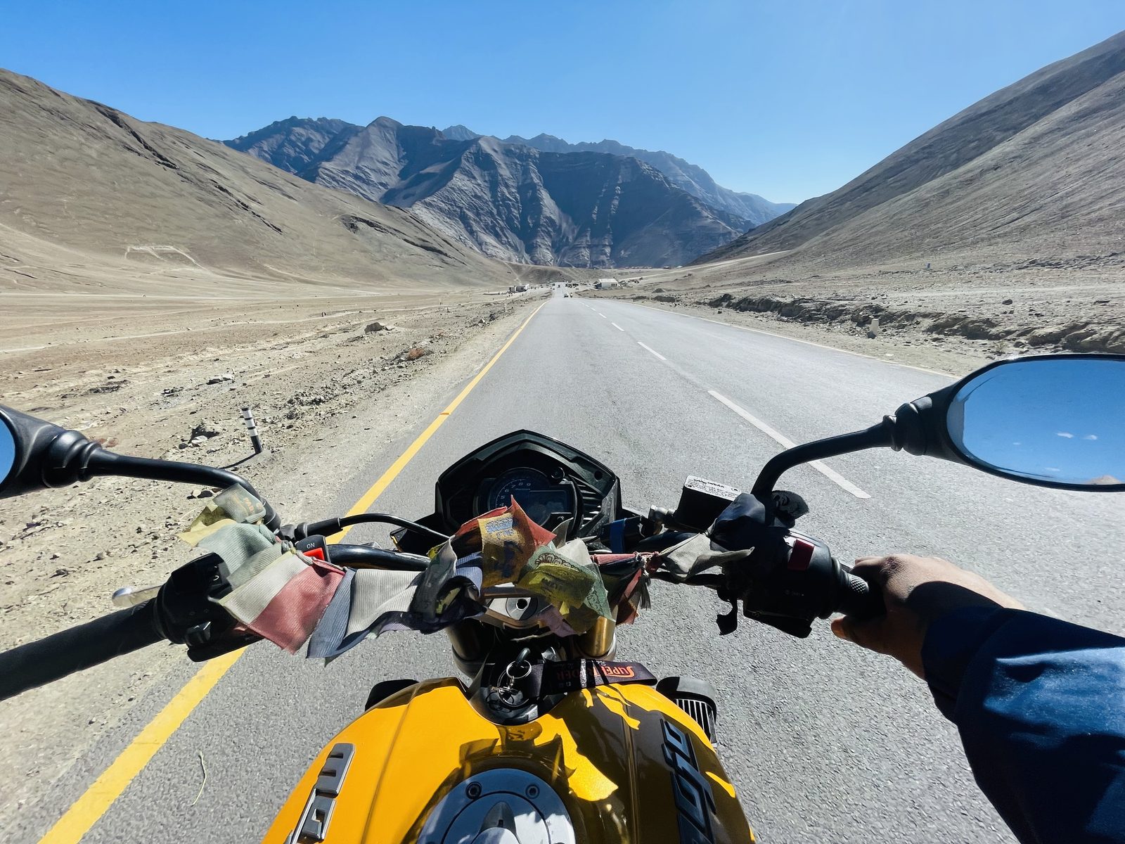 First-person motorcycle ride on the Leh Ladakh highway with prayer flags on the handlebars, open road and Himalayan mountains