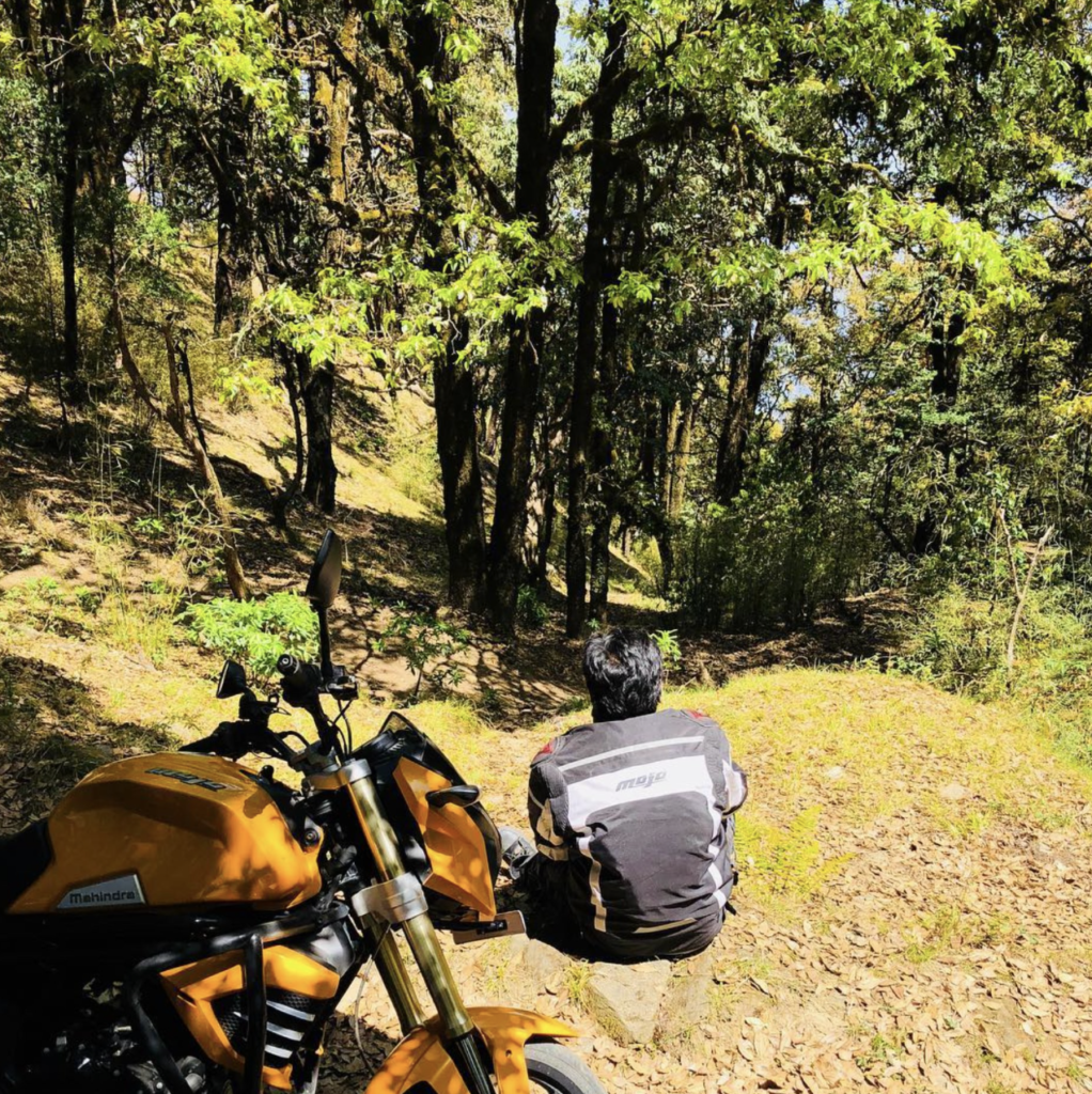 Aditya Prakash, of aadiprakash.com and Stick The Vibe, sitting on a rock beside his yellow Mahindra Mojo motorcycle on a forest trail during a Kumaon, Uttarakhand road trip.